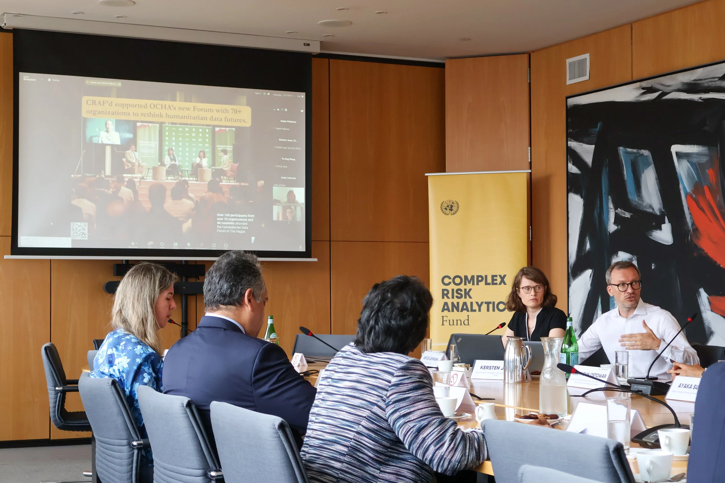 People in a conference room participating in a virtual presentation about humanitarian data, with a large screen displaying the presentation and a yellow banner for the Complex Risk Analytics Fund.