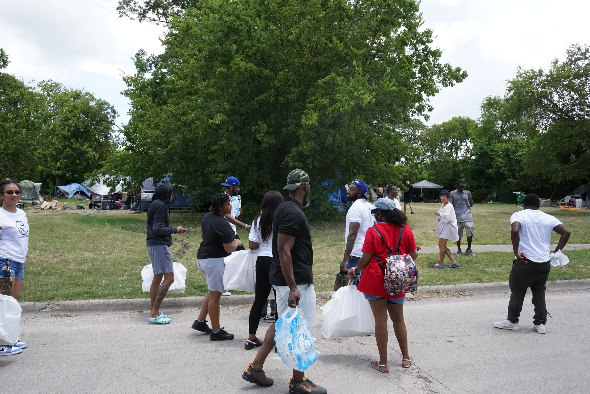 A group of people standing and walking along a sidewalk in a park or campsite area with tents in the background, surrounded by trees.