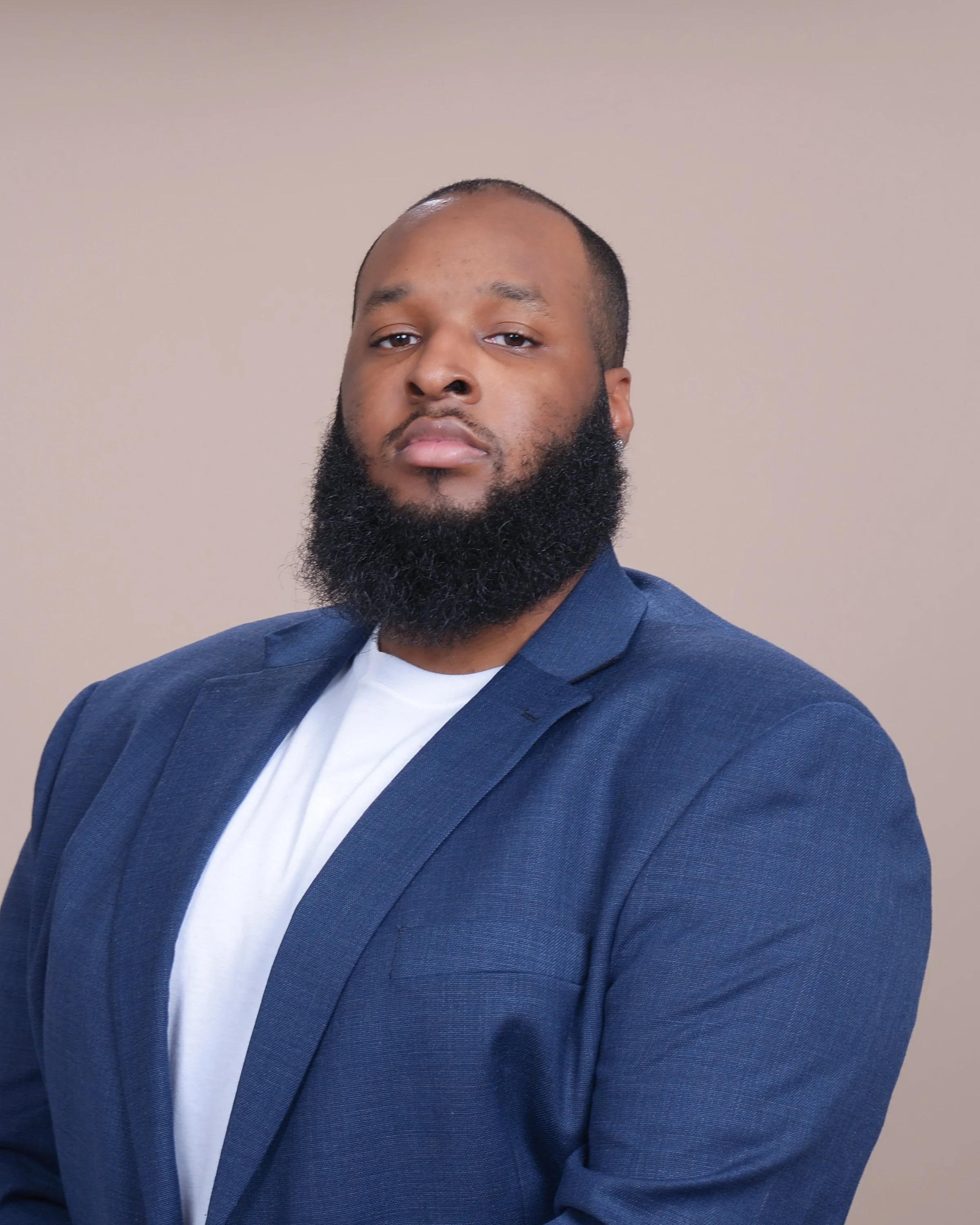 A man with a beard wearing a blue blazer and white shirt, posing against a plain beige background.