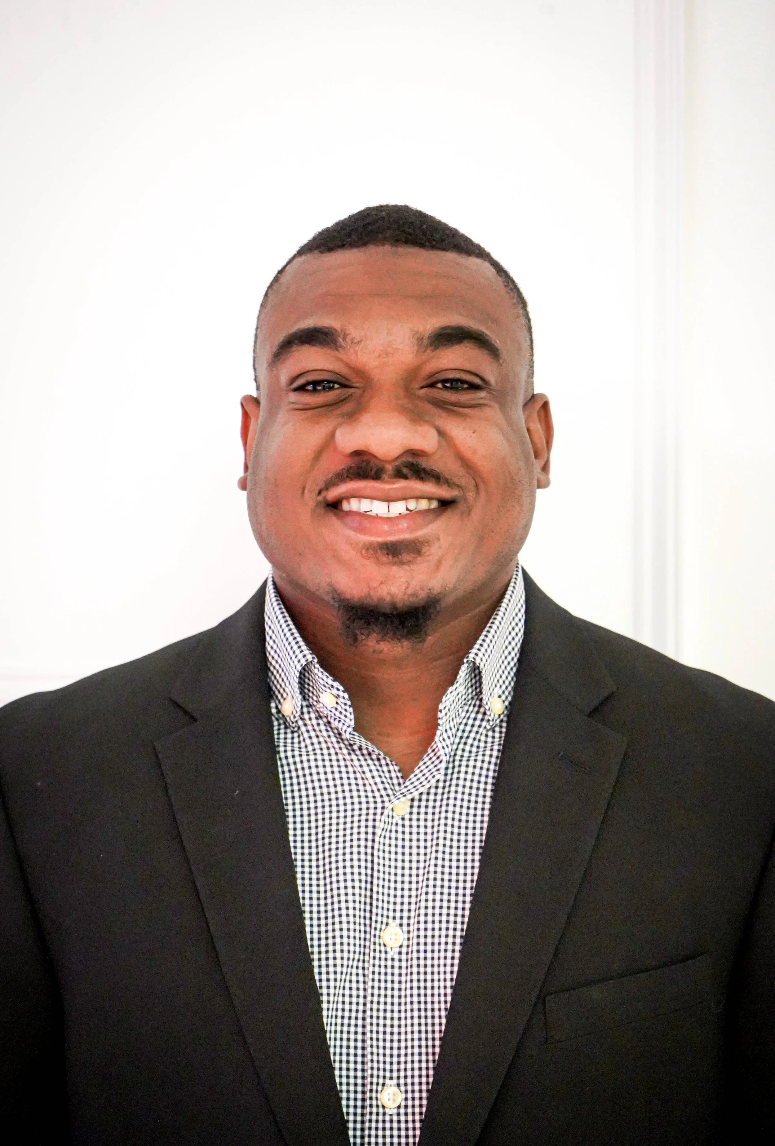 Headshot of a smiling man in a business suit and checkered shirt against a white background.