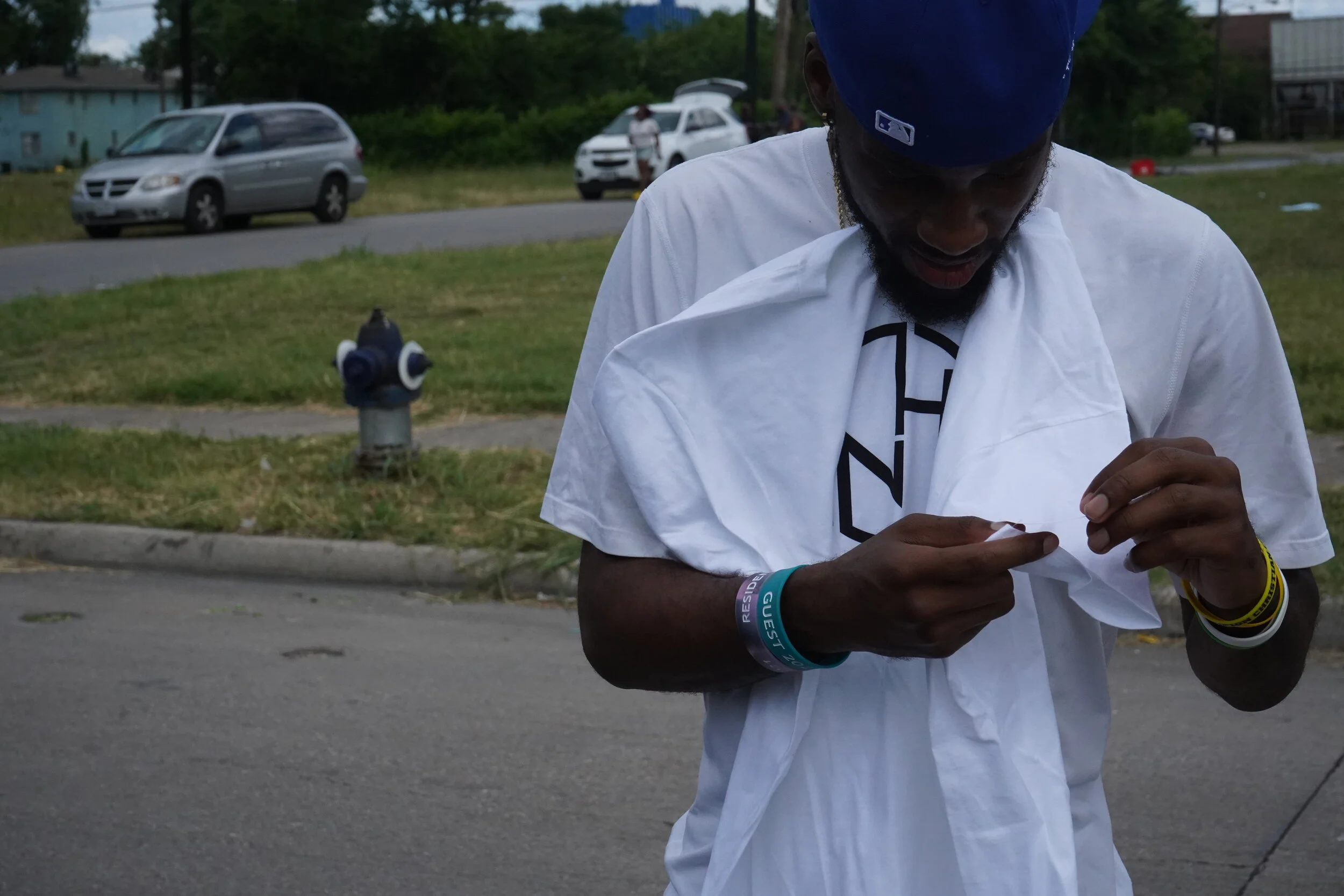 A man wearing a blue cap and white t-shirt is standing outside, holding a piece of paper and looking at it. In the background, there are parked cars, a grassy area, and a fire hydrant.