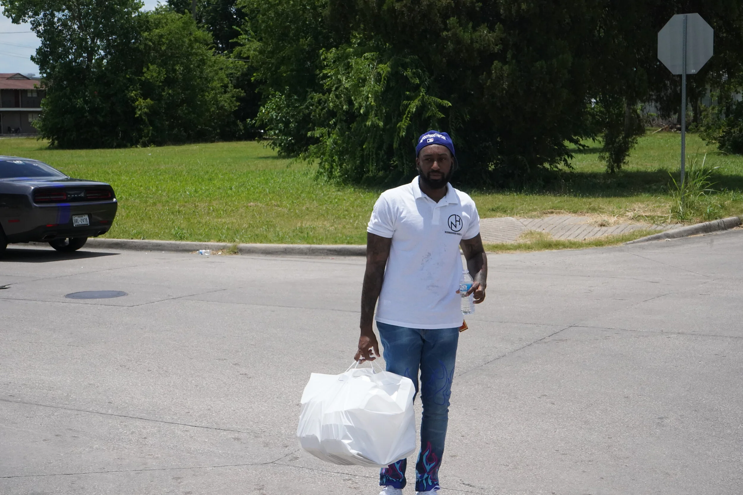 A man walking on a street holding a large white bag in his right hand and a water bottle in his left hand. He is wearing a white polo shirt, blue jeans with colorful flames on the bottom, and a blue cap. In the background, there is a black car parked