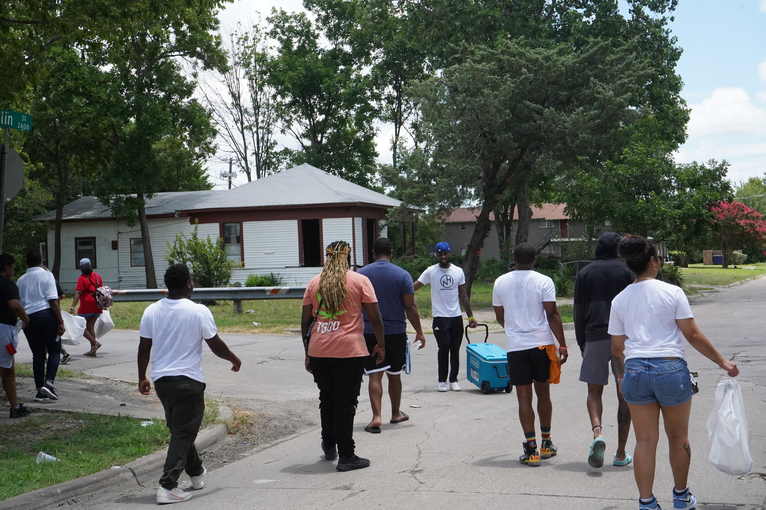 Group of people walking on a street in a residential neighborhood, some carrying bags or using coolers, with houses and trees in the background.
