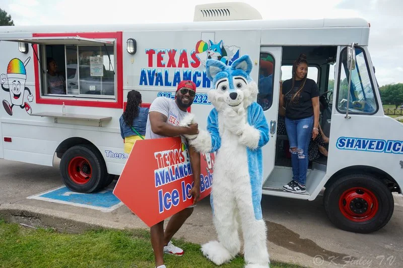 People at a Texas Avalanche Ice Ice Bar with a person in a blue and white mascot suit, and an ice cream truck in the background.