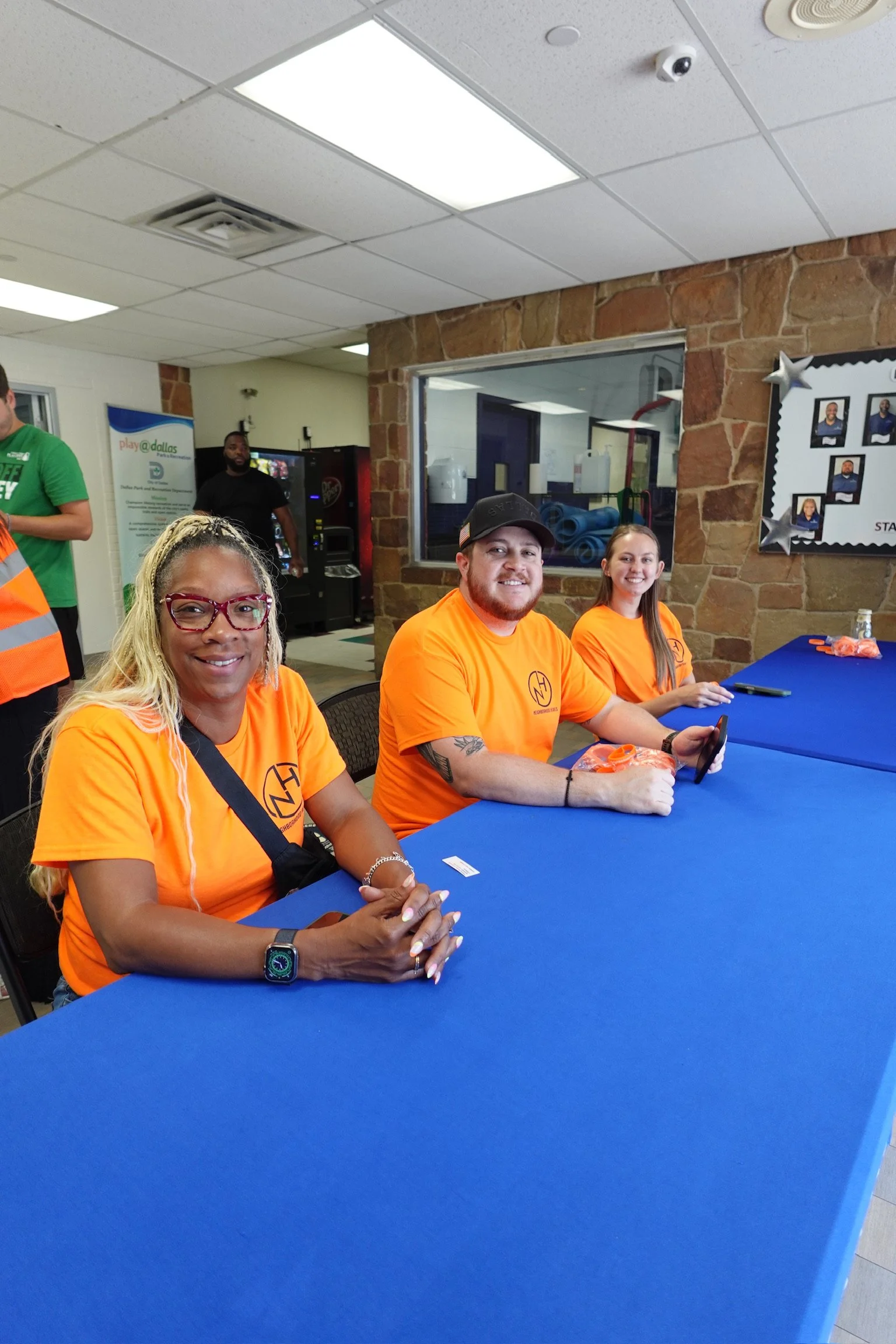 People sitting at a table wearing orange shirts, smiling, in an indoor room with a brick wall and a window.