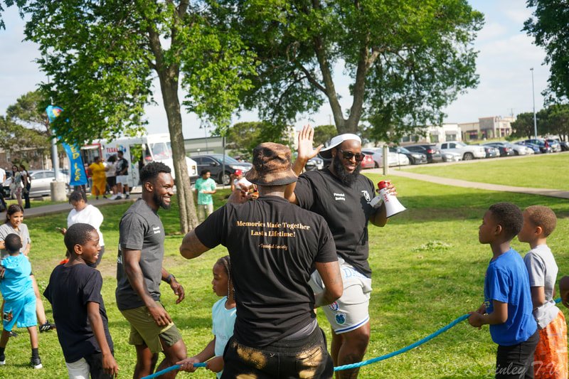 Group of people, mostly children, enjoying outdoor event in a grassy park with trees, parked cars, and tents in the background.