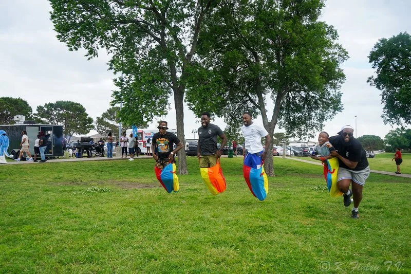 Four men playing a game with colorful bean bags on grass in a park, with trees and people in the background.