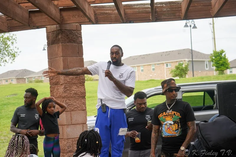 A man holding a microphone is speaking to a group outdoors under a shelter, with several people and children listening. There is a vehicle and a grassy area with houses in the background.