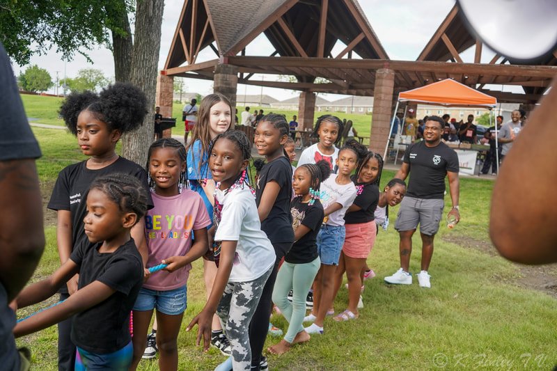 Group of children and teenagers standing in a line outdoors at a community event, with a pavilion and food tents in the background.