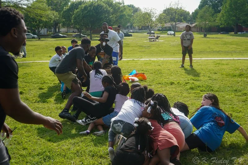 Children and adults participating in a tug-of-war game on a grassy field in a park.