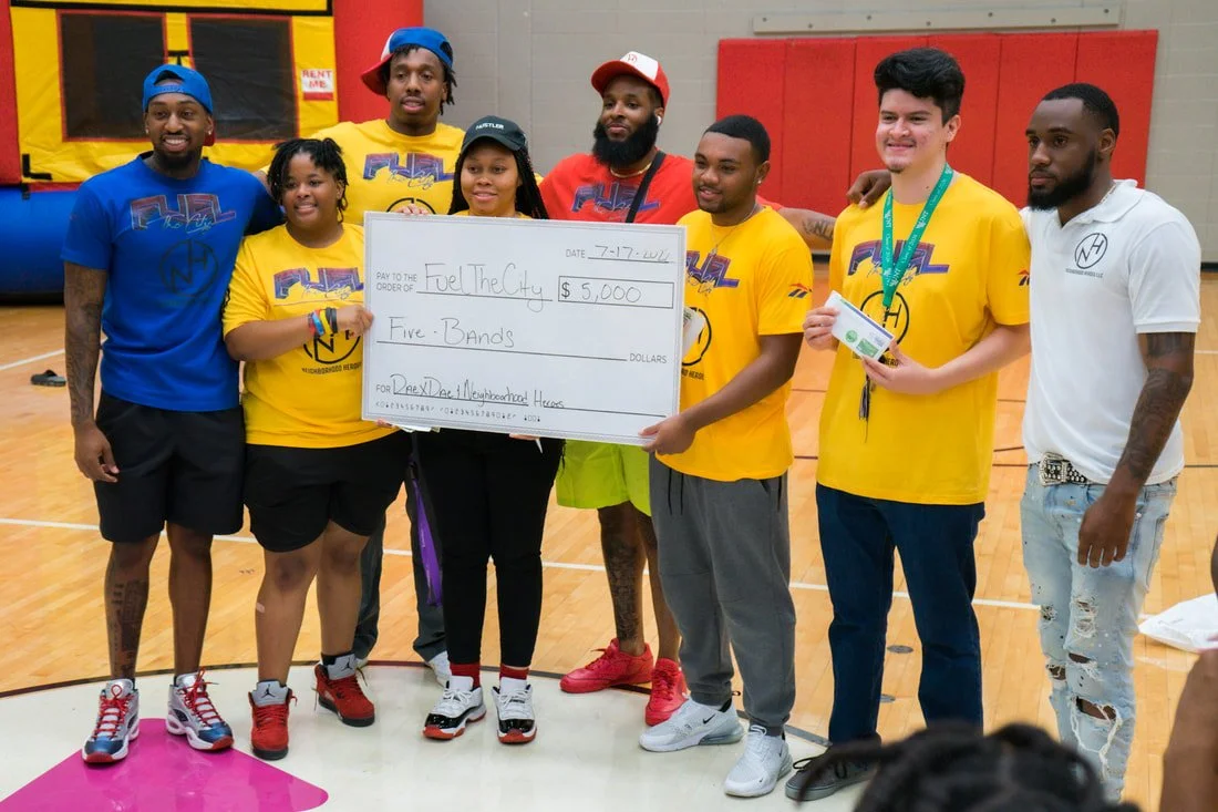 Group of seven people standing in a gymnasium, holding a large check for five thousand dollars made out to Fuel the City for a nonprofit. They are smiling and dressed casually, some wearing matching yellow T-shirts with a logo, and wearing medals or caps.