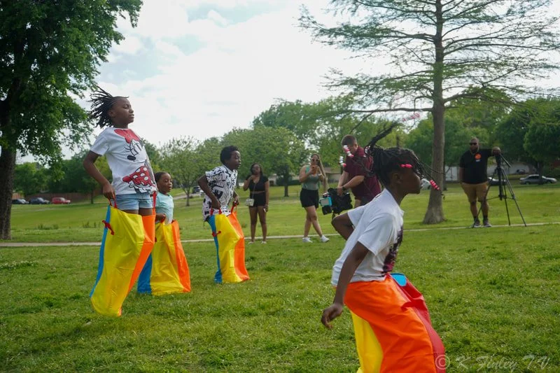 Children playing sack race outdoors in a park with onlookers and trees.