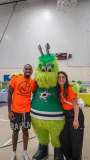 A person in a green bunny mascot costume with big eyes and long ears, wearing a Dallas Cowboys jersey, standing between two young adults, one on each side, all inside a gymnasium.
