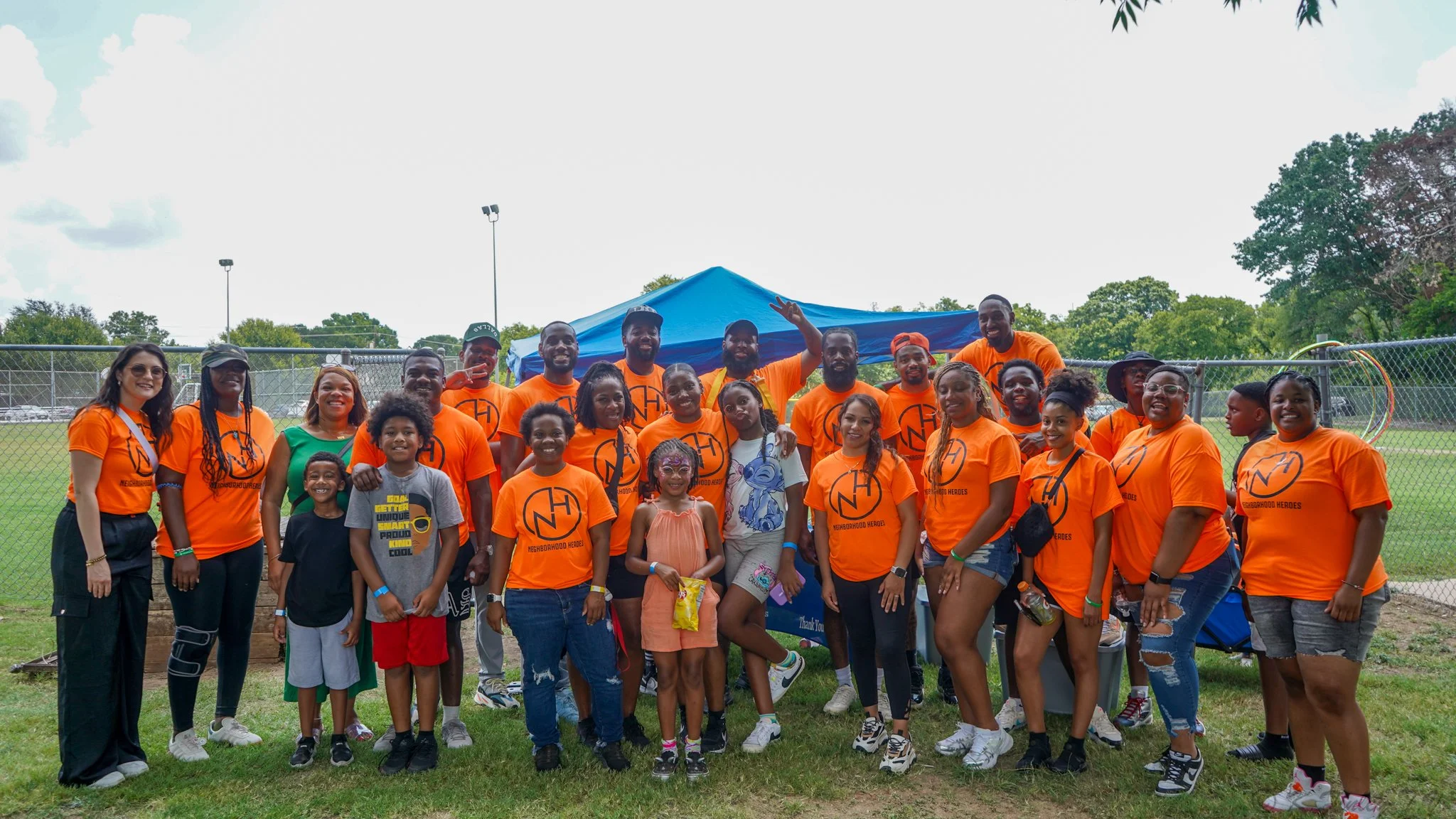 A group of people, including children and adults, standing outdoors on grass in front of a chain-link fence, smiling for a photo. Many are wearing bright orange T-shirts with a logo and text, and there is a blue canopy tent behind them.