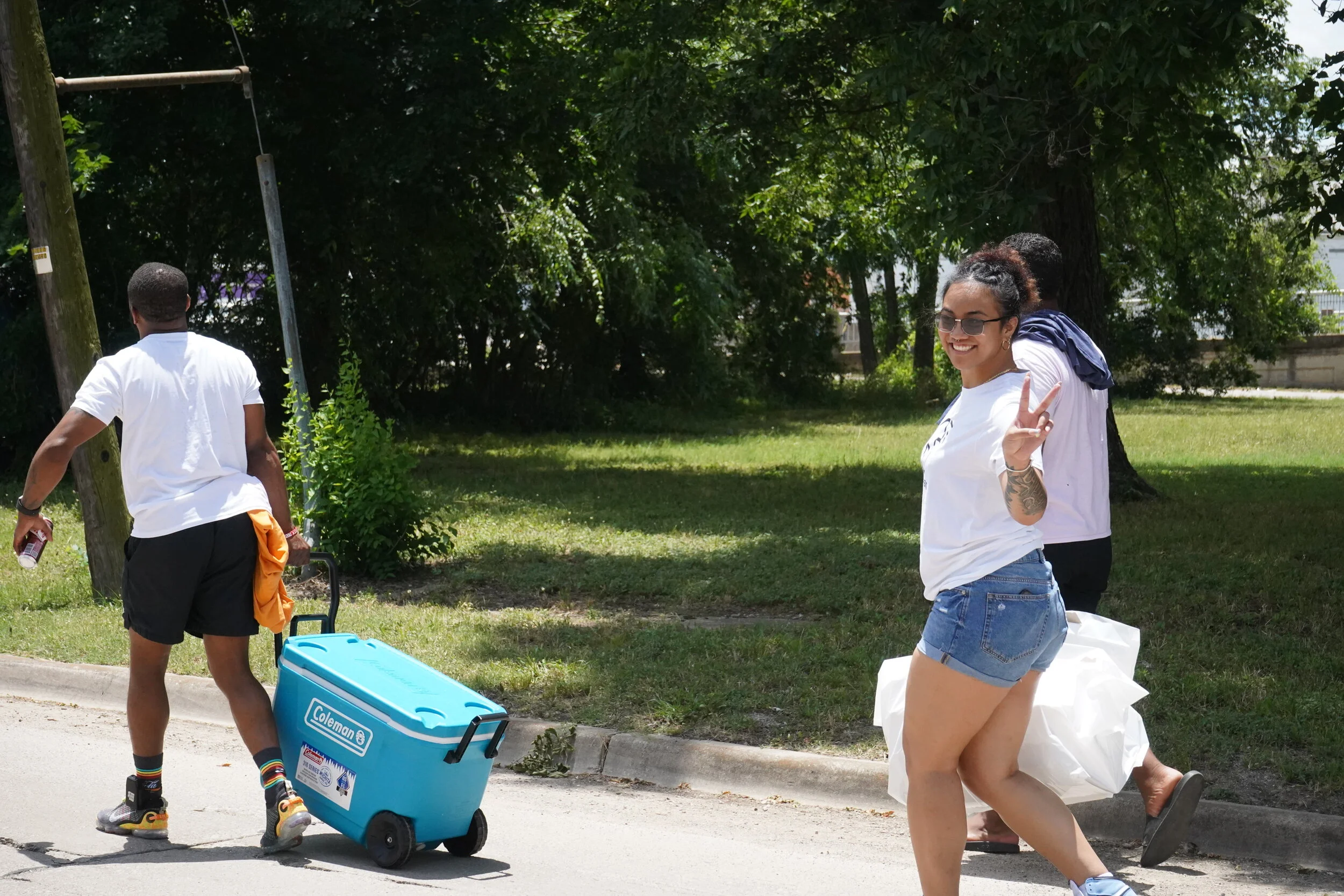 A smiling young woman with glasses and a tattoo on her arm is walking along a sidewalk, giving a peace sign. She is wearing a white t-shirt and denim shorts. In the background, a person with a backpack and white t-shirt is walking on the grass, and a