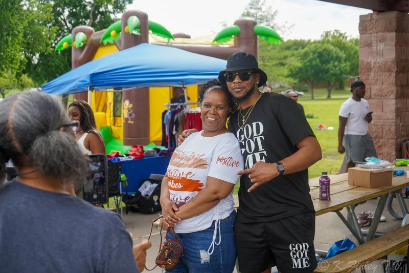 A woman and a man pose for a photo at an outdoor event, with a bounce house and various tables in the background.