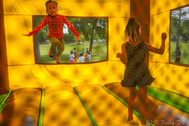 Two young girls are playing inside an inflatable bounce house; one girl is jumping near the window, while the other is standing on the bounce surface.