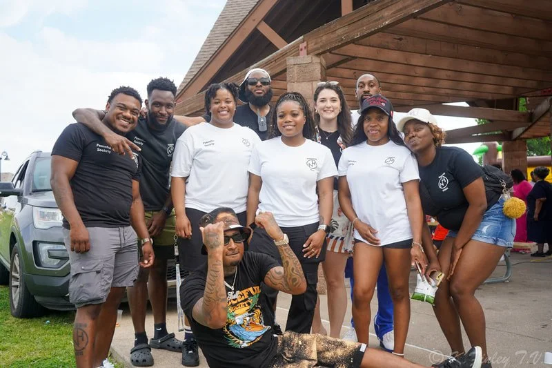 Group of nine diverse people posing outdoors at a social event, some standing and one sitting in front, under a wooden pavilion with a vehicle and other attendees in the background.