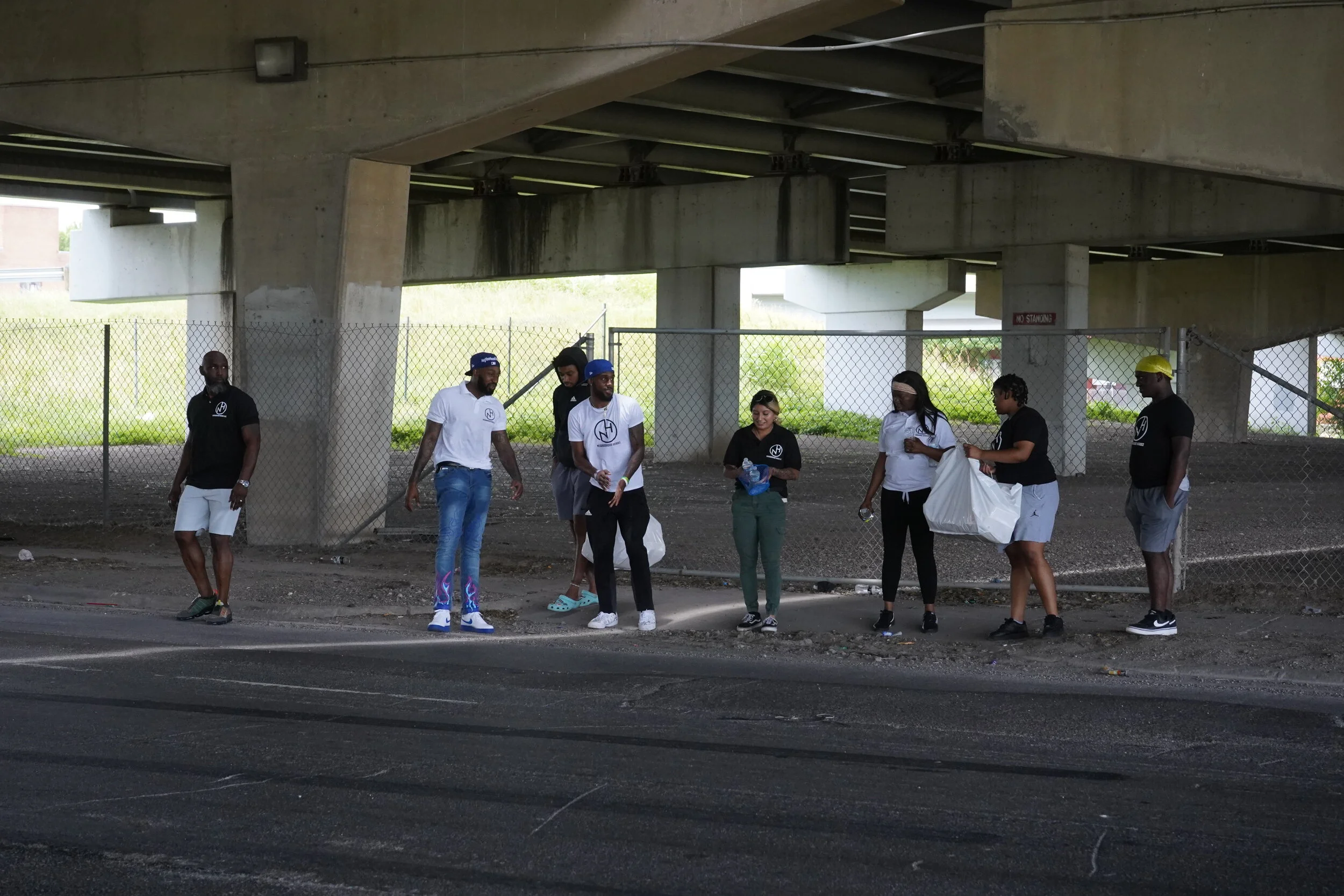 A group of people standing under a highway overpass, some holding bags and water bottles. They are engaged in conversation or looking around, with a chain-link fence behind them and patches of grass visible through the fence.