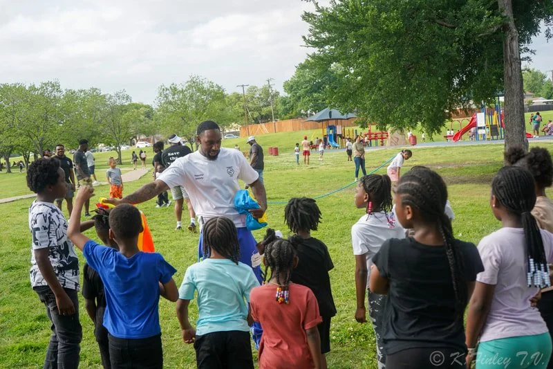A man in a white shirt distributes water balloons to children at an outdoor park with a playground in the background.
