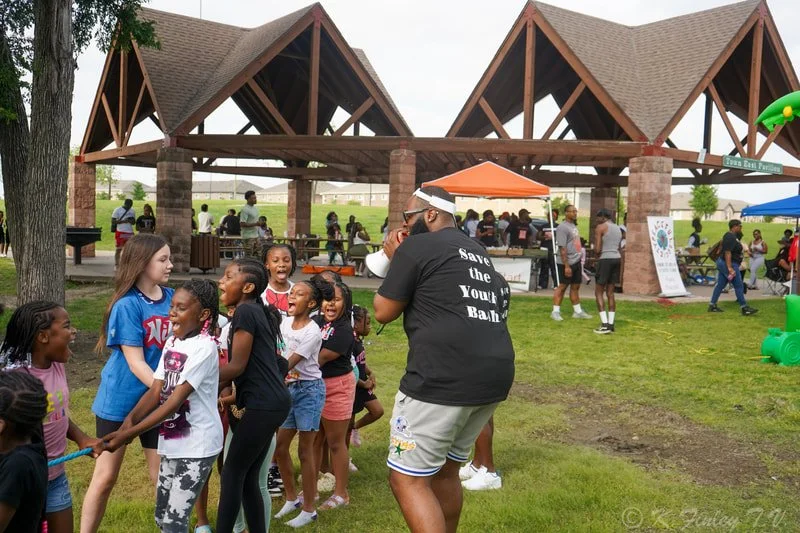 Children playing tug-of-war at an outdoor event with a pavilion and tents in the background.