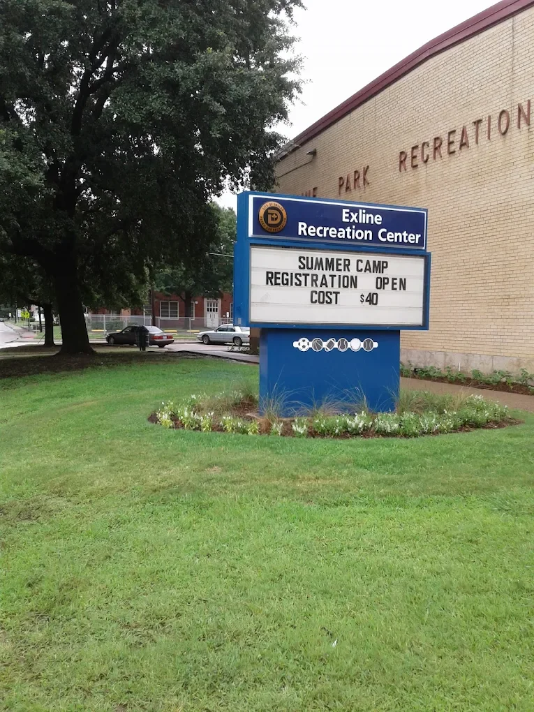 Sign for Exline Recreation Center advertising summer camp registration, open now, costing $40, on a grassy area with flowers, with a brick building and parking lot with cars and trees in the background.