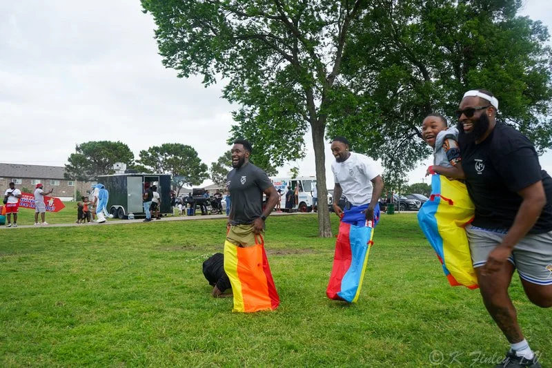 Group of people playing sack race outdoors at a community gathering or event, with a woman dressed as a clown in the background.