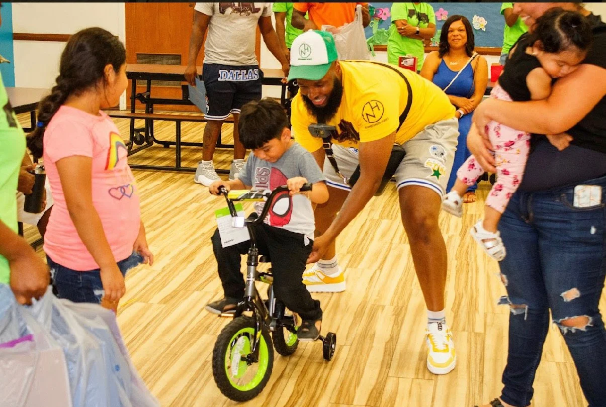 A young boy learning to ride a small bicycle with training wheels, guided by an adult man, in a room with other children and adults watching and smiling.