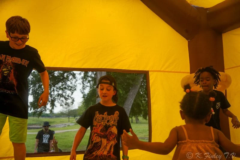 Four children playing inside a yellow tent. One girl with pigtails and two boys are visible, all engaging with each other, with a screened window showing an outdoor scene in the background.