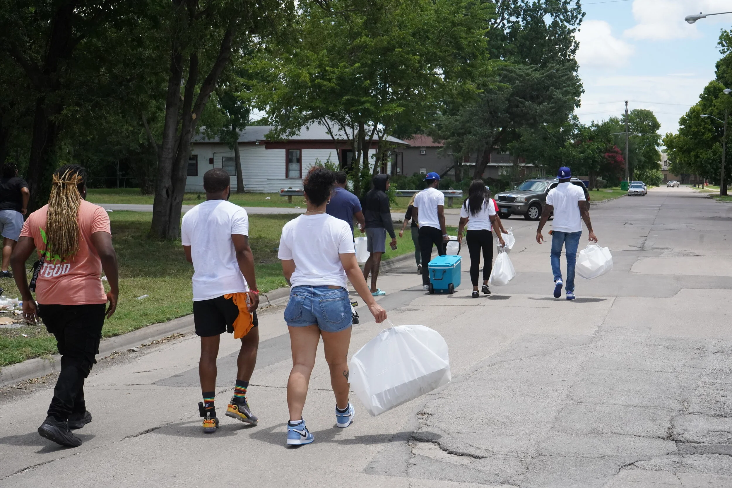 Group of people walking on a street carrying bags and supplies, with trees and houses in the background.