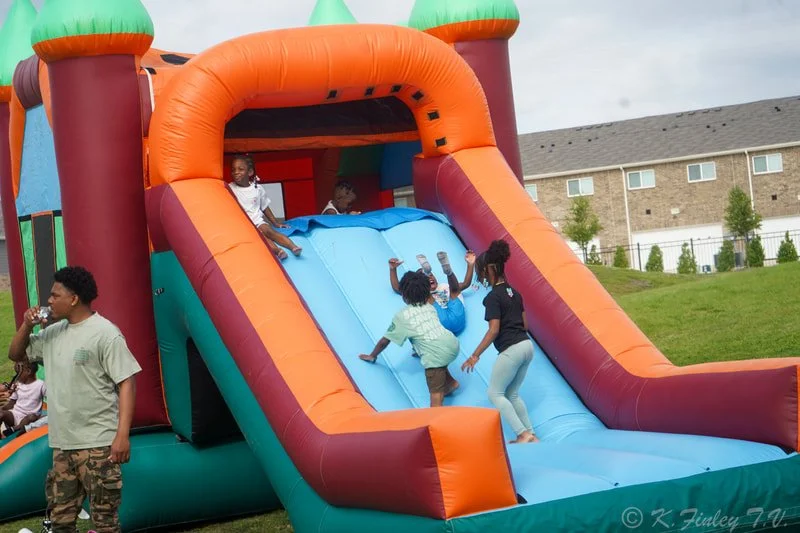 Kids playing on a colorful inflatable slide at an outdoor event, with some climbing up and others sliding down, while an adult stands nearby.