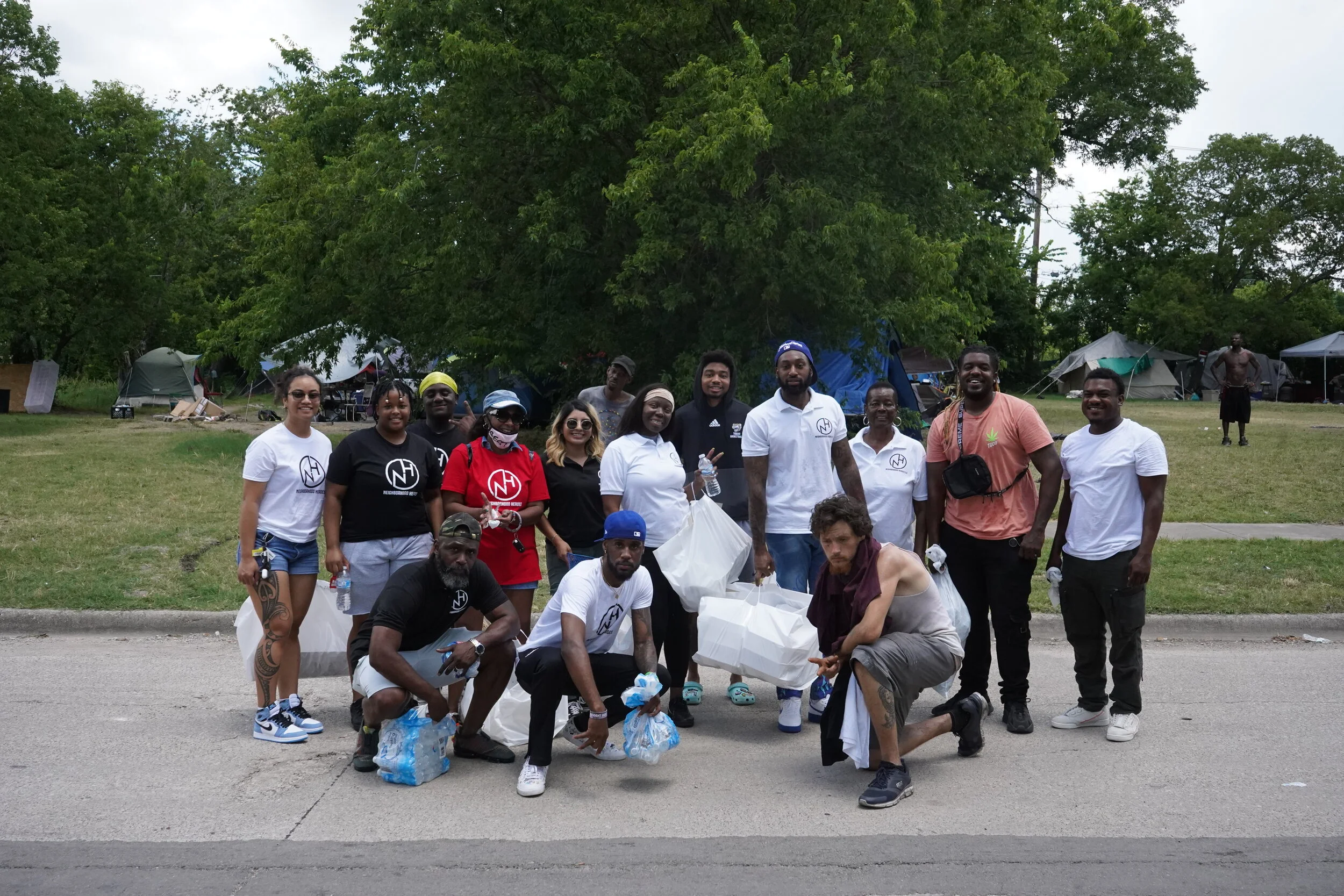 Group of people outdoors standing and kneeling in front of tents, holding trash bags and water bottles after a cleanup event.