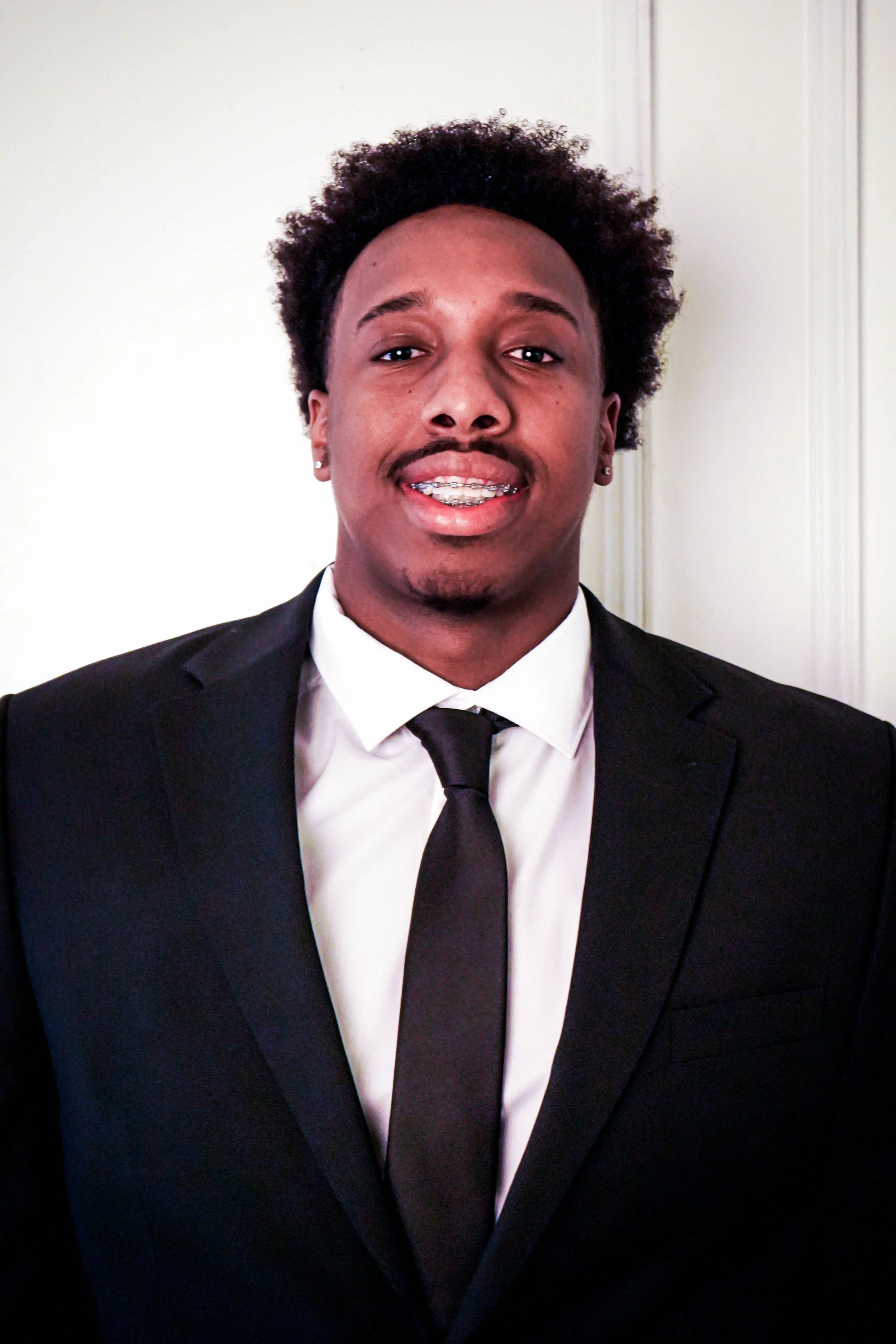 Young man in a black suit, white shirt, and black tie smiling in front of a neutral background.