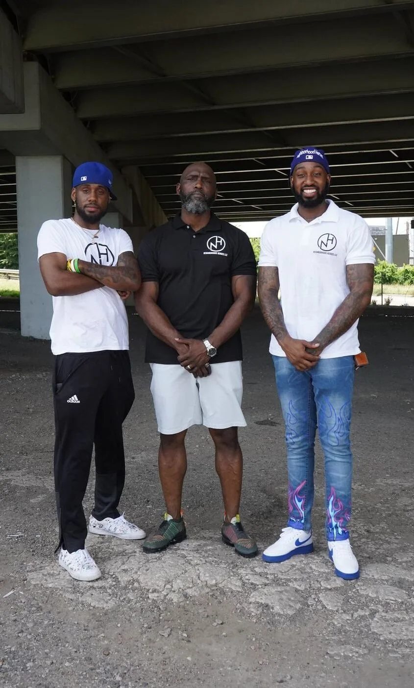 Three men standing under a bridge outdoors, posing for a photo. The man on the left is wearing a white t-shirt and black pants, the man in the middle is wearing a black polo shirt and shorts, and the man on the right is wearing a white polo shirt and blue jeans with colorful patterns.