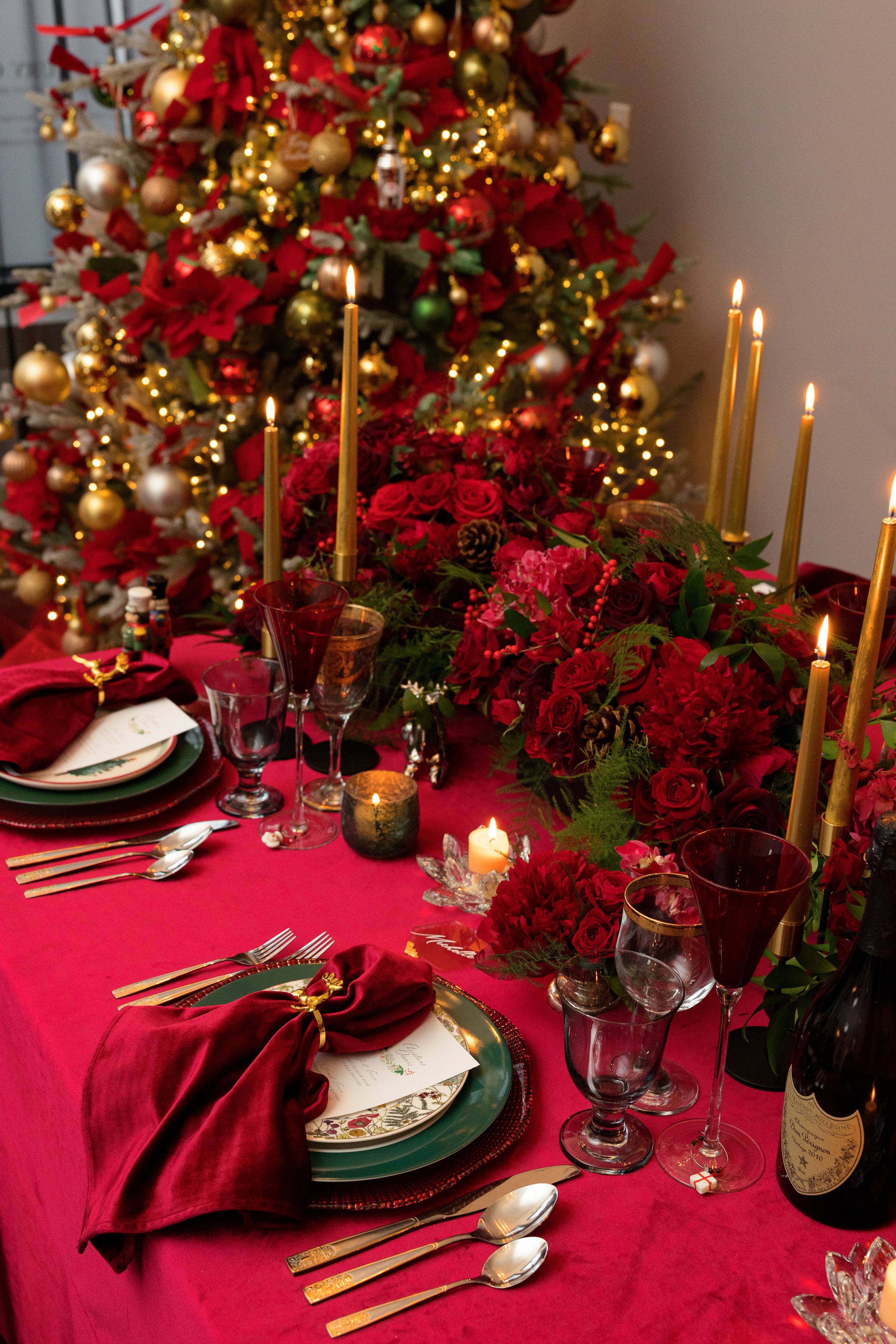 Holiday dinner table with red roses, gold taper candles, layered place settings and a decorated Christmas tree