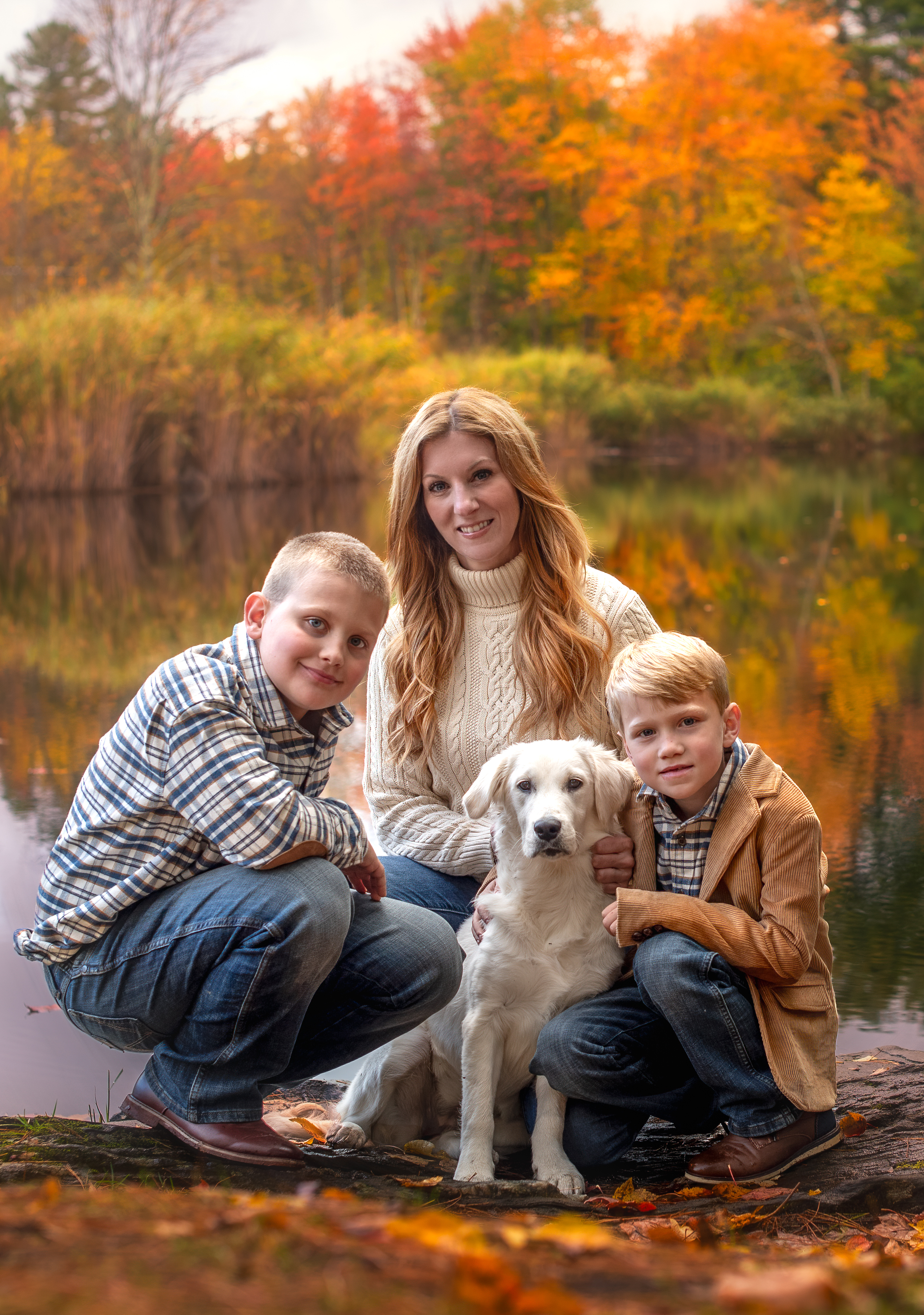 Meghan and her two sons and dog smile in front of an autumn lake scene