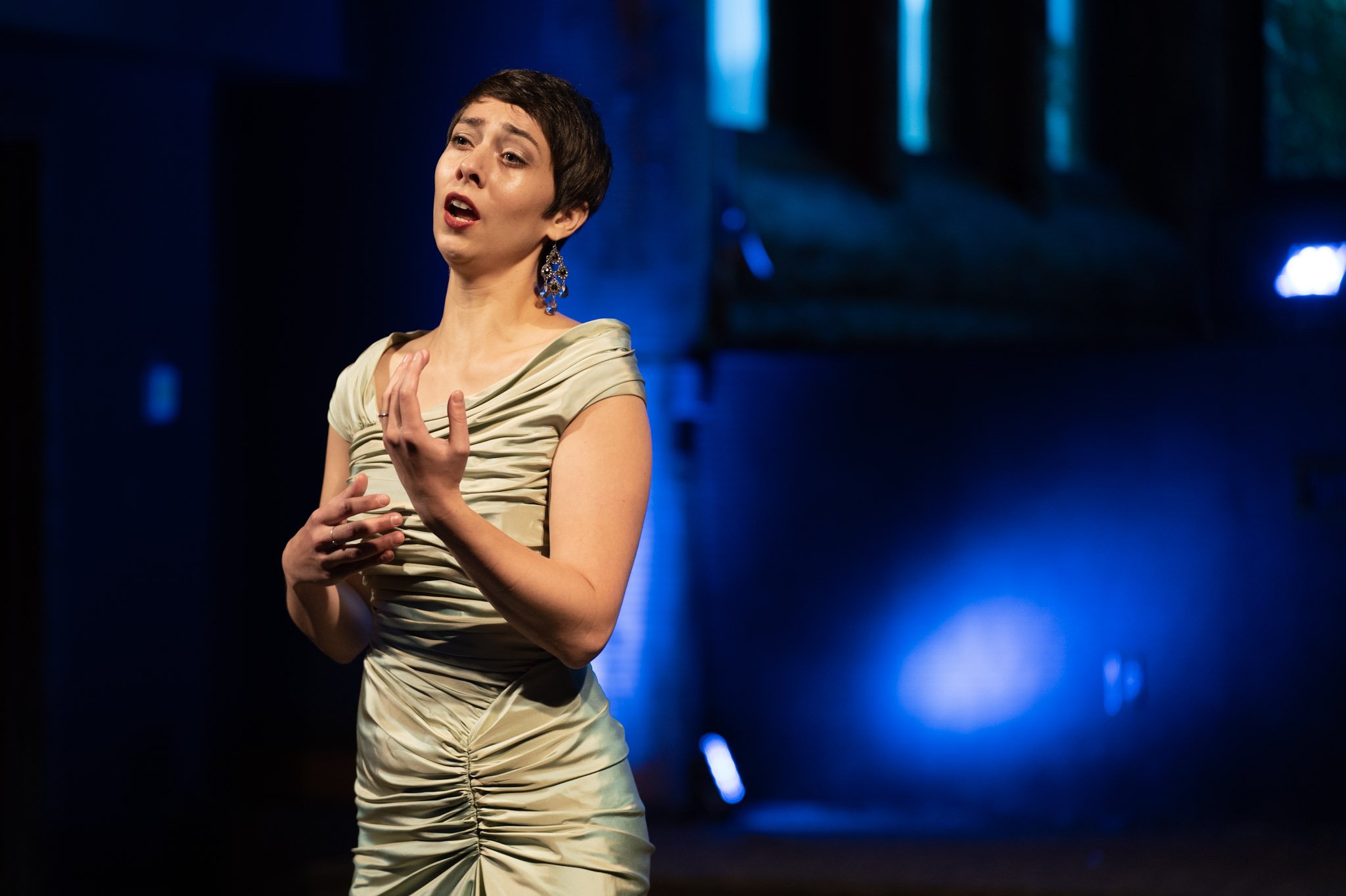 Female opera singer performing on stage with expressive gestures, wearing an elegant green dress, illuminated by blue and purple stage lighting.