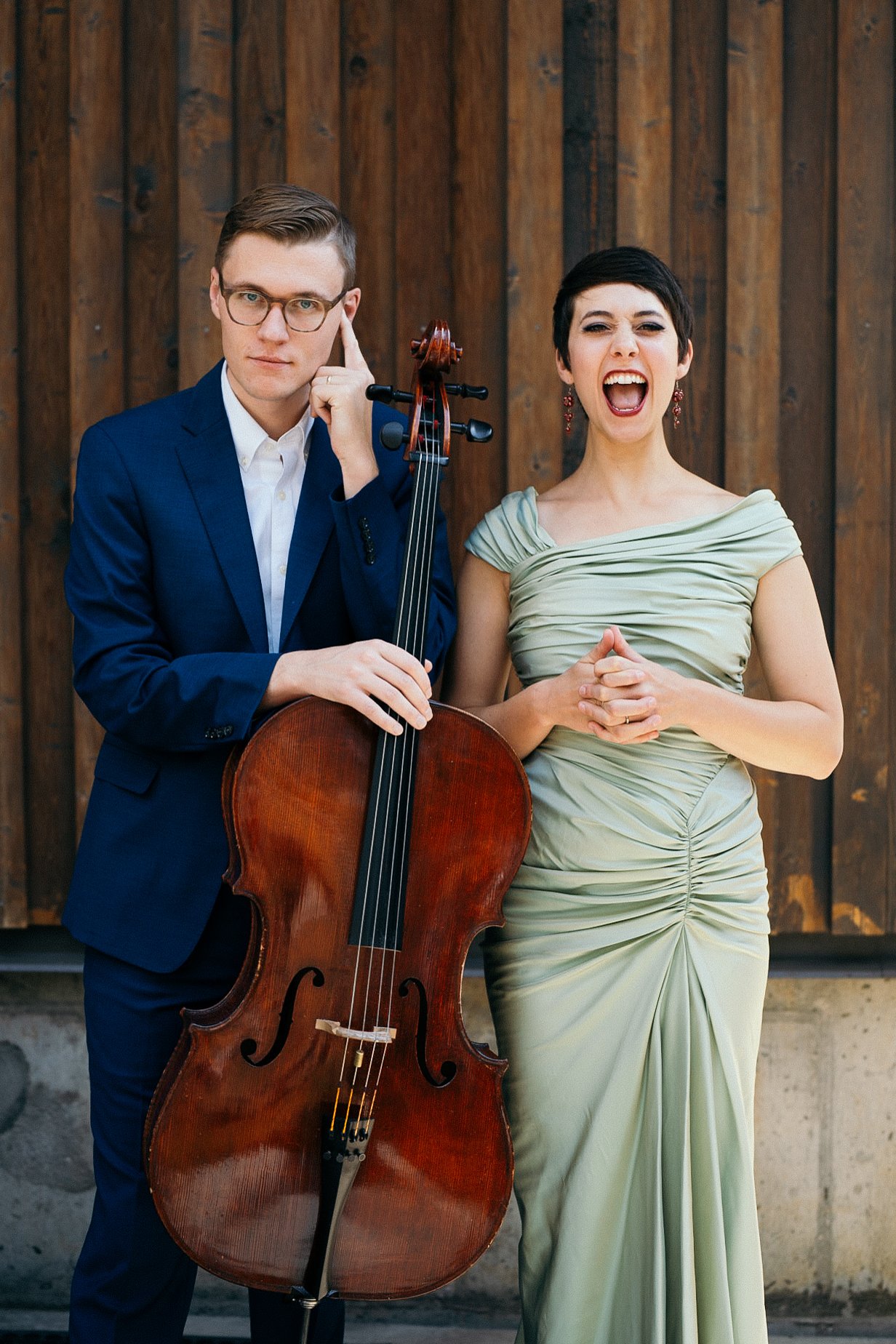 Man in blue suit holding a cello, standing next to a woman in a light green dress, both posing in front of a wooden wall.