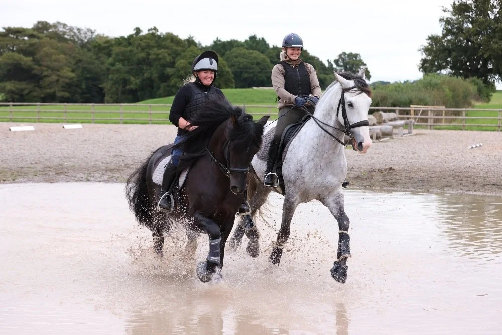 Two smiling riders riding their horses, centred in the middle of a water splash obstacle. A chunky black pony powers through with a dappled grey elegant horse to its left.