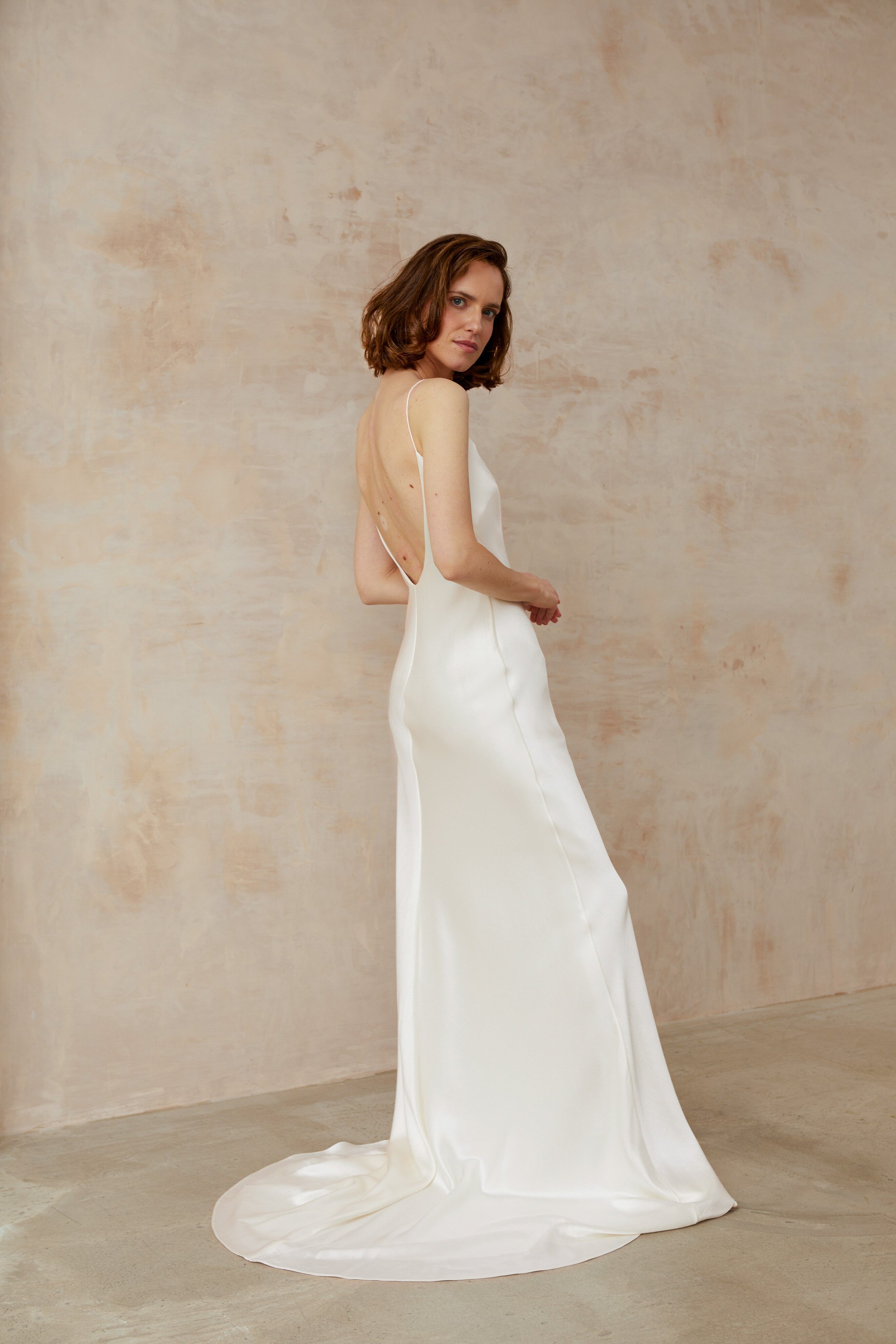 a woman wearing a ivory dress, standing poised in front of a wall. This exquisite minimal square neck bias slip style wedding dress showcases a captivating low back, epitomizing timeless beauty.