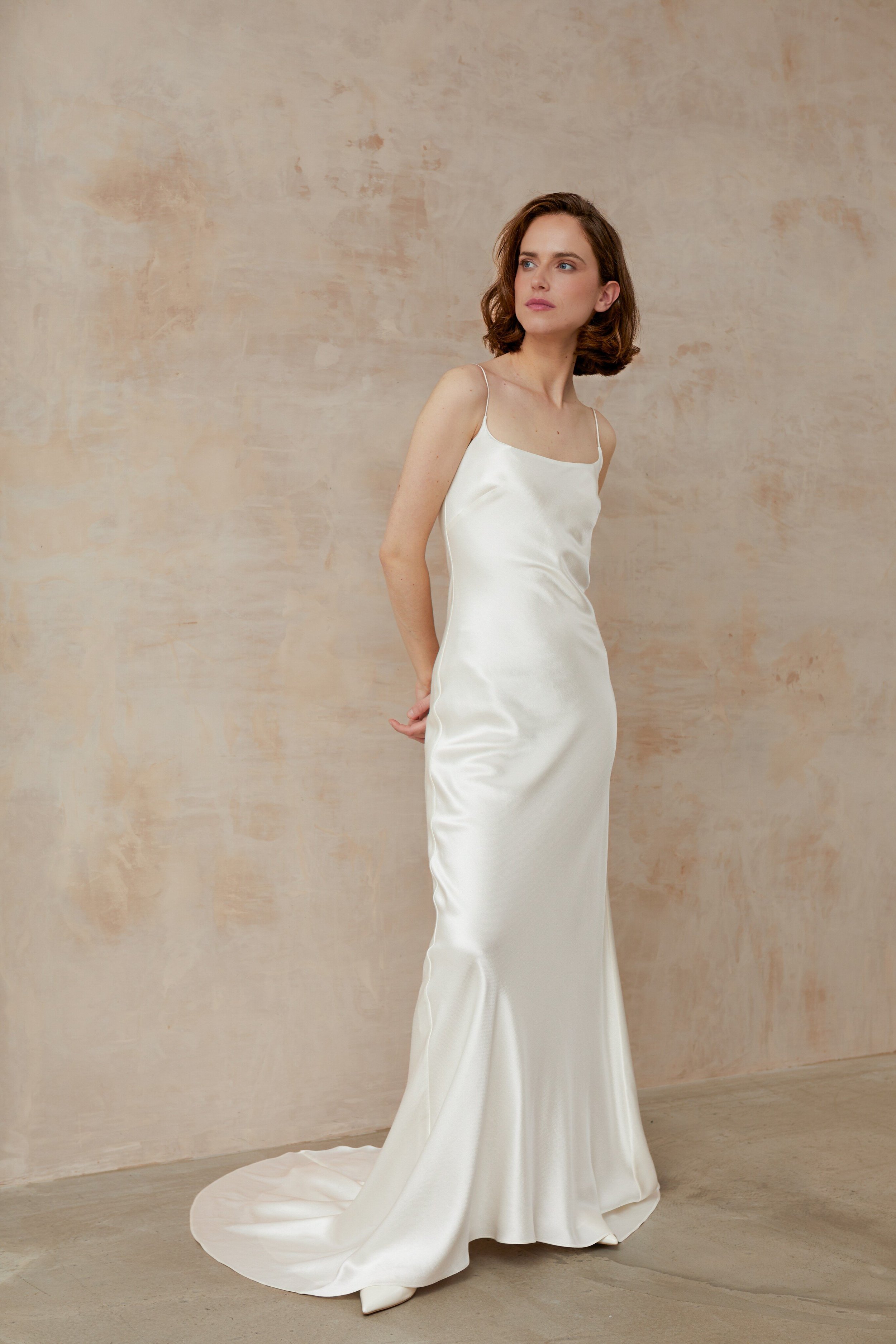 Elegant woman in a minimal square neck wedding dress standing gracefully in front of a nude wall.