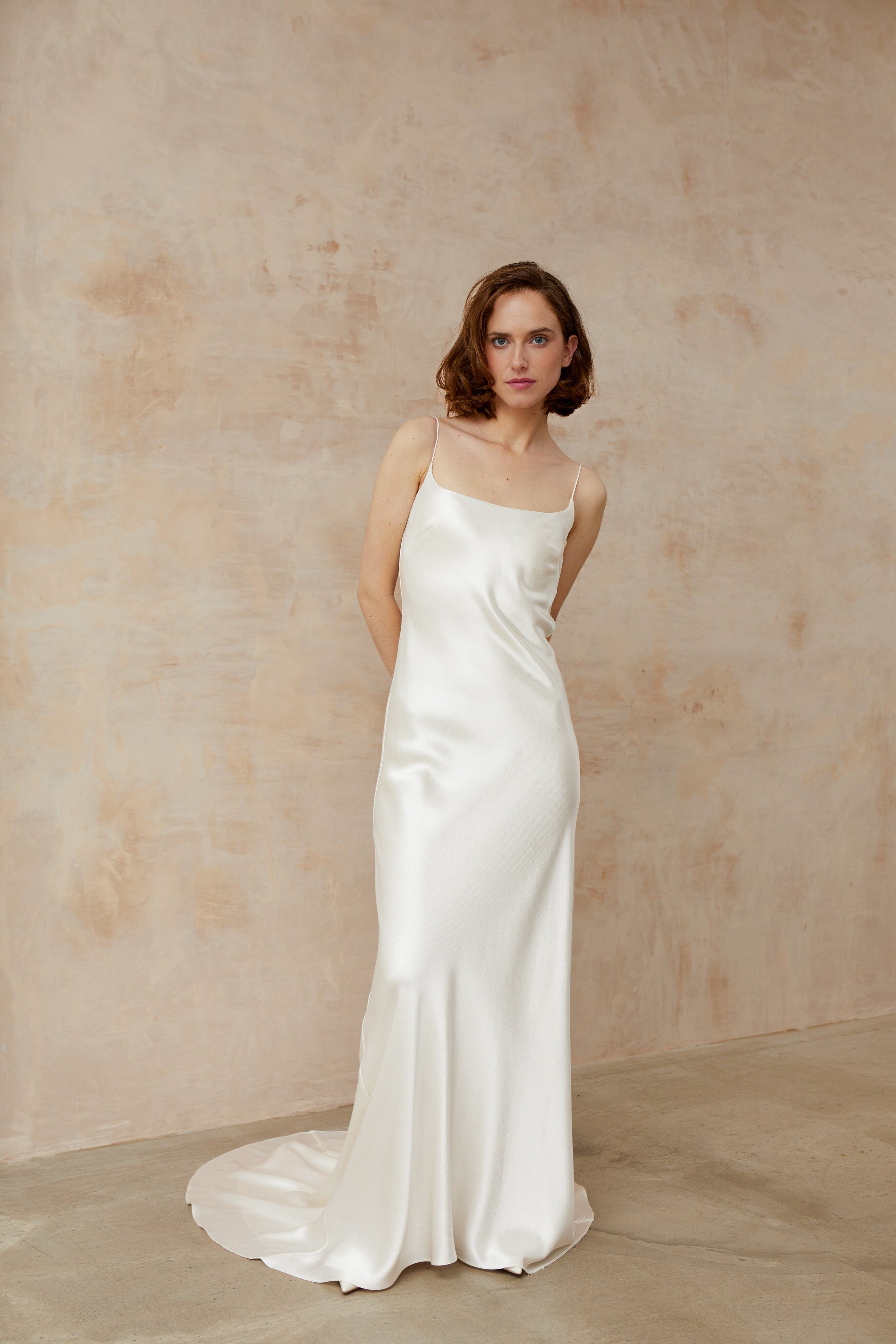 Graceful lady in a white dress with square neckline stands in front of a stylish wall.
