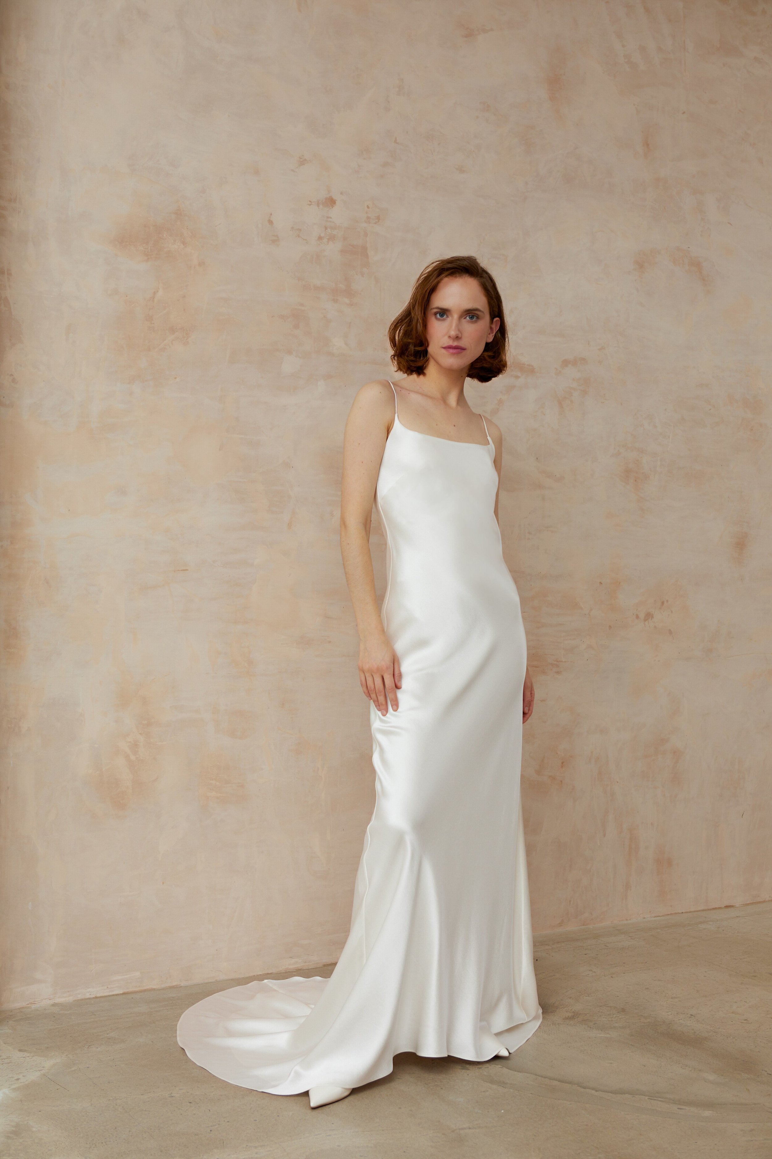 Graceful lady in a white square neck wedding gown posing in front of a clean nude wall.