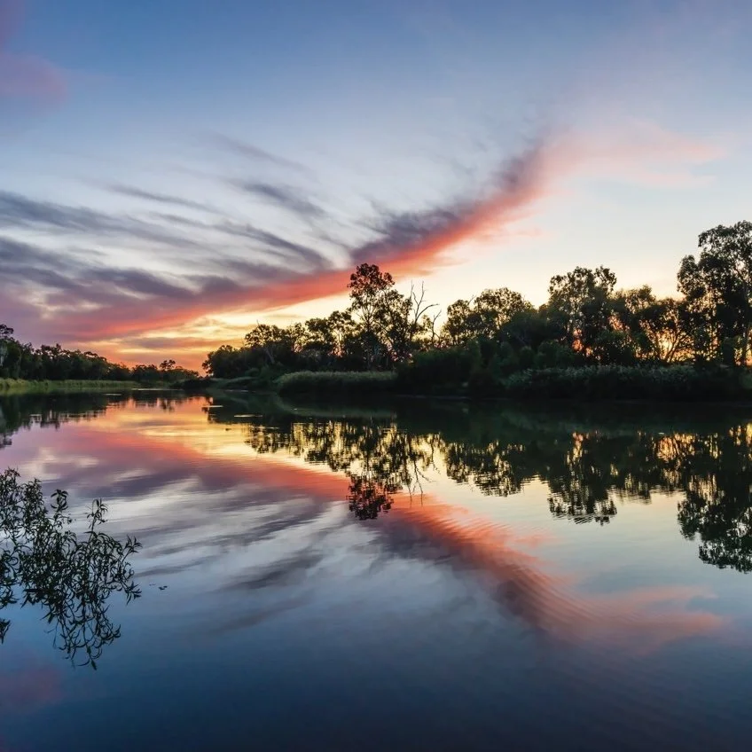 A tranquil river reflects the vibrant colors of a sunset, with hues of pink, orange, and purple streaking across the sky. Silhouettes of trees line the riverbank, creating a peaceful and picturesque natural scene. The still water mirrors the sky’s colors, adding to the serene atmosphere of the moment.