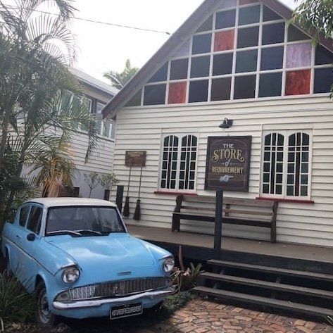A white wooden building with a sign that reads “The Store of Requirement.” The building has arched windows and stained glass near the roof. In front, a light blue vintage car is parked partly in the garden. The setting is surrounded by tropical plants.