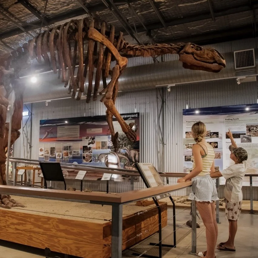 A large dinosaur skeleton exhibit is displayed in a museum with informational panels in the background. A woman and a child stand nearby, observing the fossilized remains.