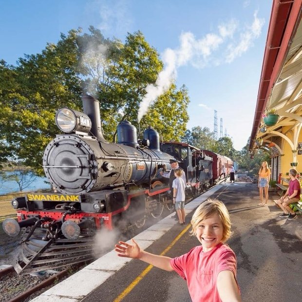 A smiling child in a red shirt stands on a train platform, taking a selfie with a black steam locomotive labeled “Swanbank” in the background. Steam rises from the engine, and people are seen boarding or admiring the train.