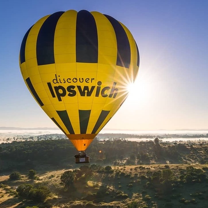 A bright yellow and black hot air balloon with “Discover Ipswich” written on it gracefully floating over a scenic landscape at sunrise.