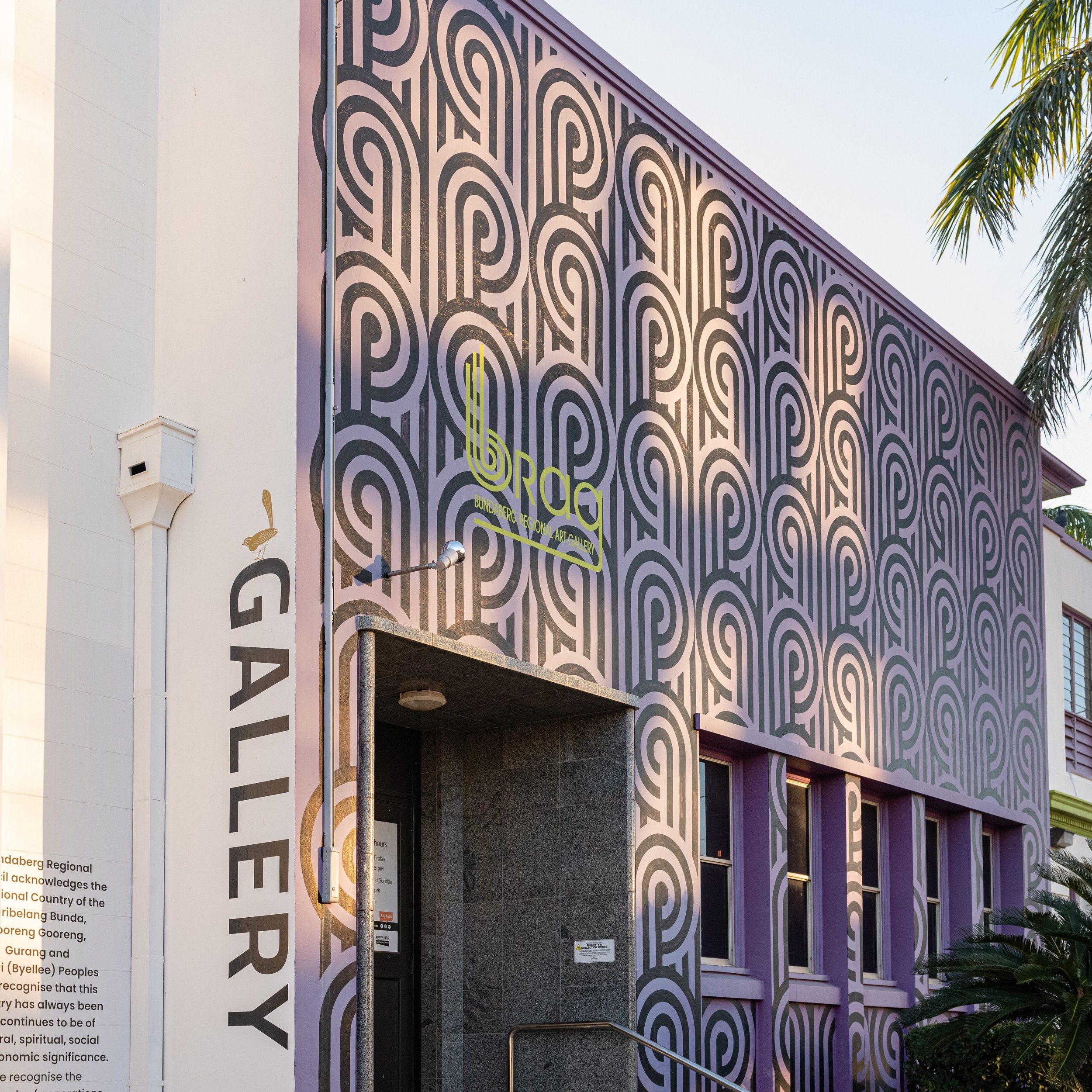 Exterior view of the Bundaberg Regional Art Gallery, featuring a bold geometric mural with black and pink patterns on the building facade.