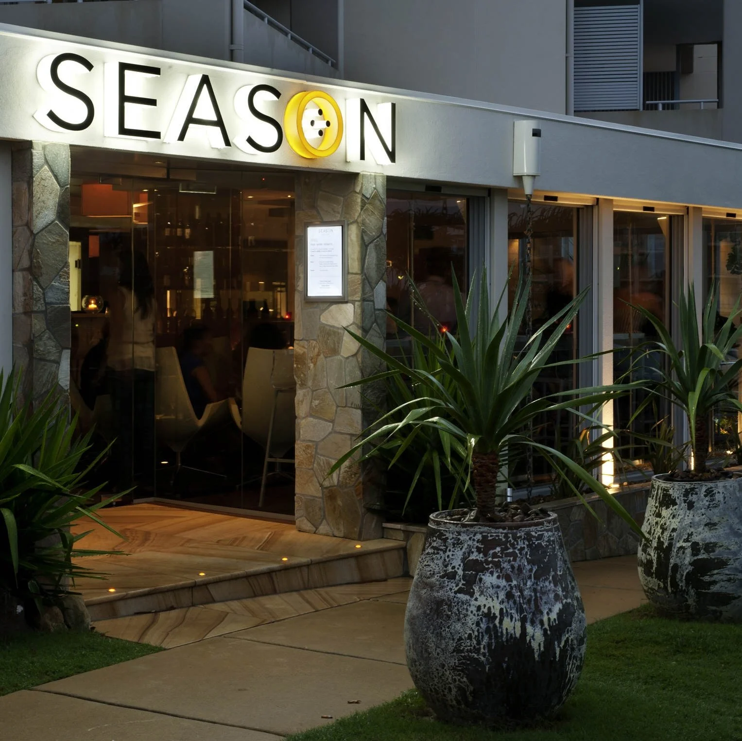 Exterior of Season Restaurant at dusk, with large potted plants flanking the entrance. The glass doors reveal a warmly lit interior with seated patrons, and the restaurant’s sign features the word “SEASON” with a stylized yellow “O” resembling a sun or abstract design.
